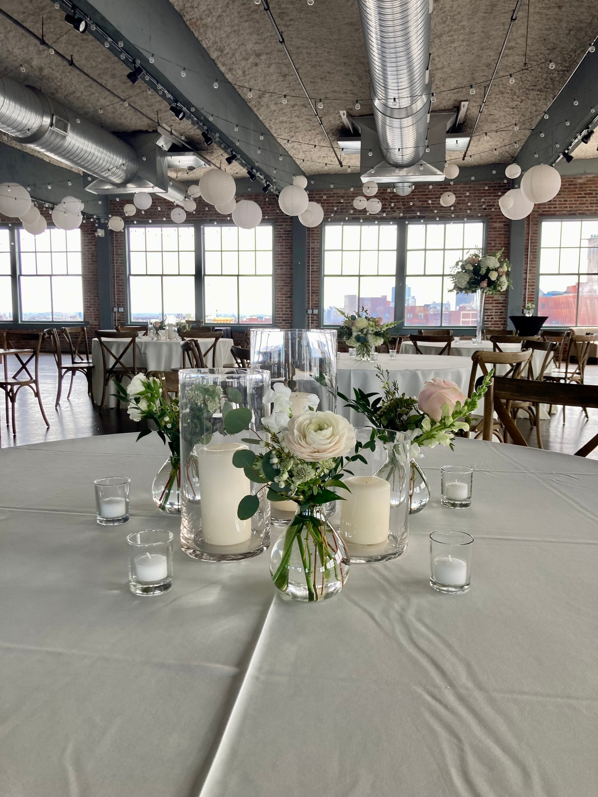 Decorated tables with white floral centerpieces and candles in a large room with high ceilings and large windows.