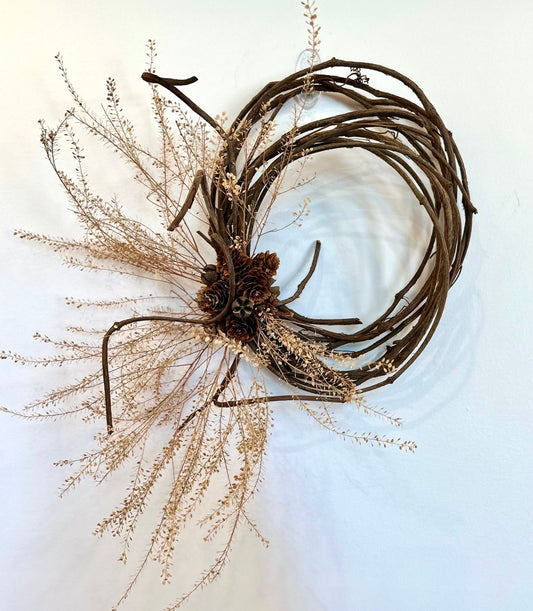 Penelope Wreath: a white wall with a dried wreath made of grapevine and adorned with pennycress and pinecones