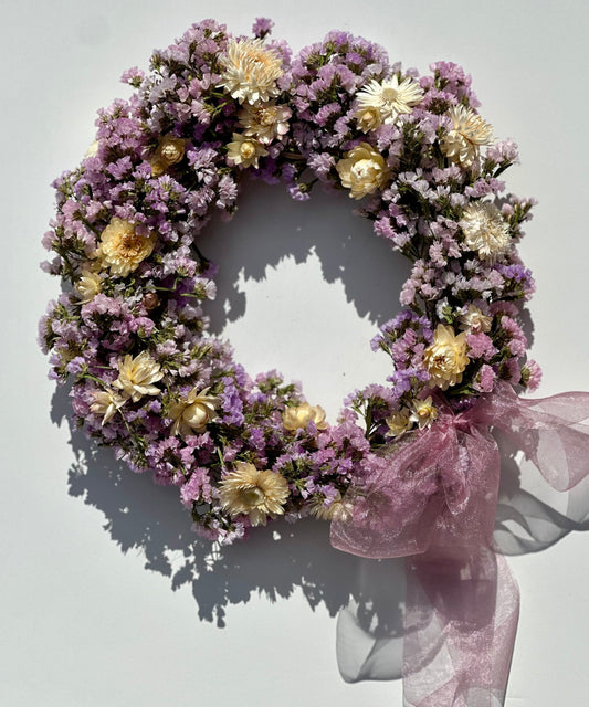 Eloise Wreath: a white wall with a wreath of dried lavender statice and dried buttercream strawflower adorned with a gauzy lavendar bow