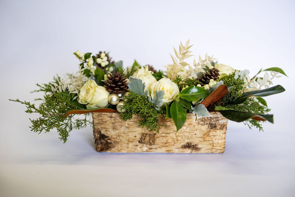 White floral arrangement with pinecones and greenery in a wooden flower box on a light background