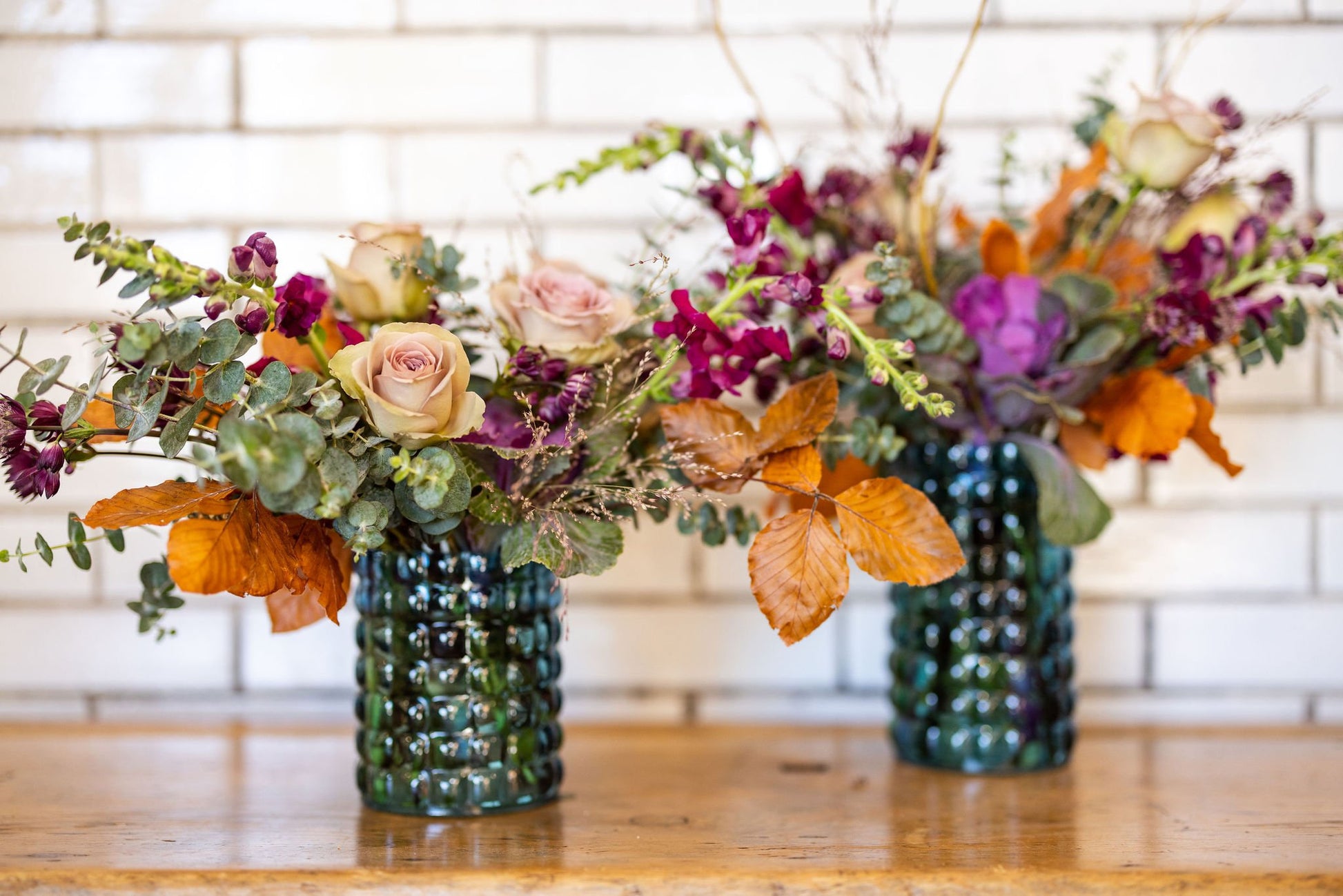Two colorful flower arrangements of oranges and magenta in green vases on a wooden surface with a blurred white background.