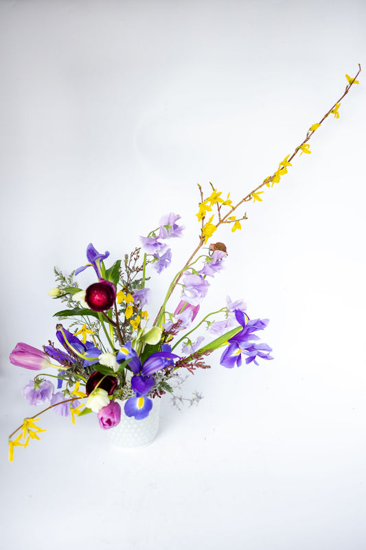 A white vase featuring a floral arrangement of vibrant and colorful blooms in shades of purple, lavender, mauve, white, sunshine yellow, and deep burgundy.