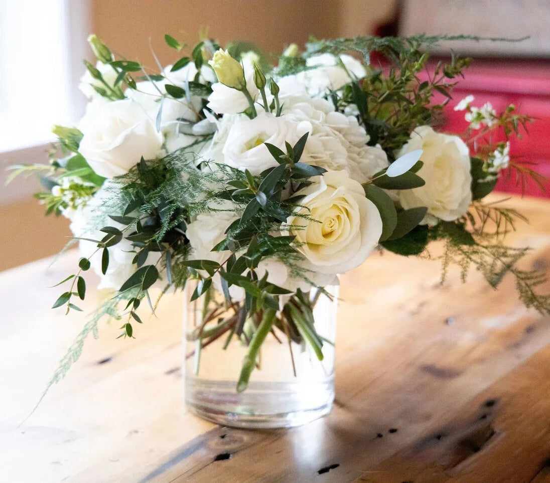 Bouquet of white flowers and greenery in a clear vase on a wooden surface.