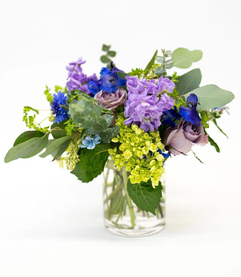 Bouquet of various blue and lavender flowers in a clear vase on a white background