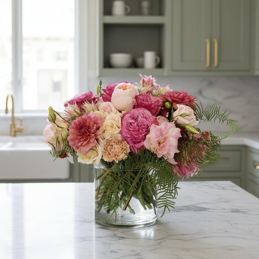 Bouquet of pink, blush, and cream flowers in a clear vase on a light background in a kitchen
