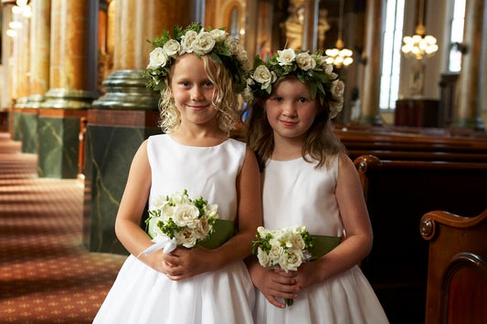 Two young girls in white dresses with floral crowns standing in a church.