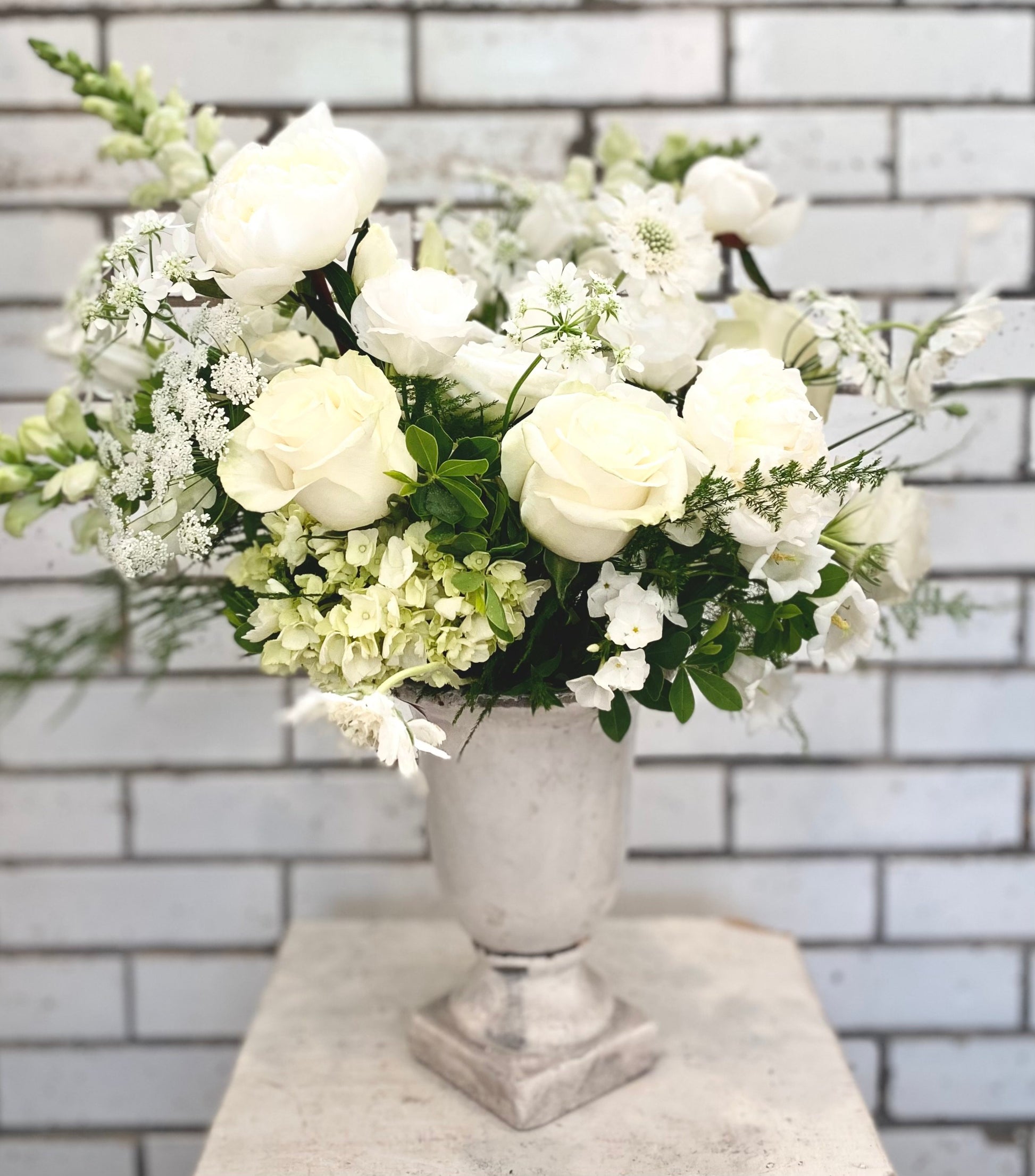 Bouquet of white flowers in a stone vase against a white brick wall.
