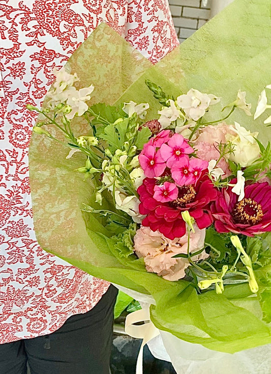 Bouquet of bright pink, blush, and white flowers wrapped in green paper with a person wearing a red and white patterned shirt in the background.